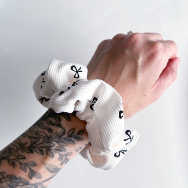 A white scrunchie with a ribbed texture and small black bow-tie shapes is worn on a wrist with a tattoo'd arm.